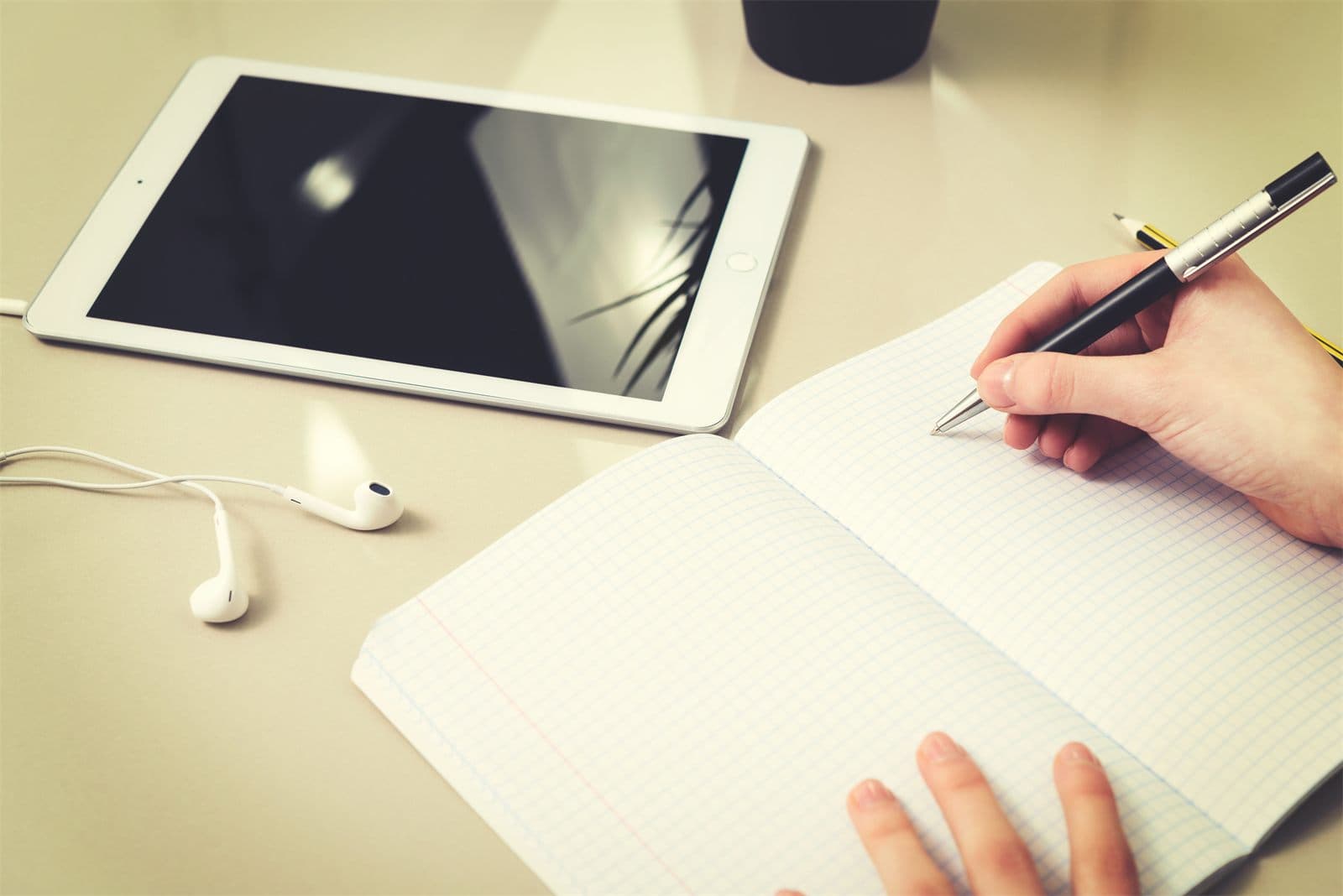 Une main qui écrit sur un cahier, à côté d'une tablette posée sur un bureau